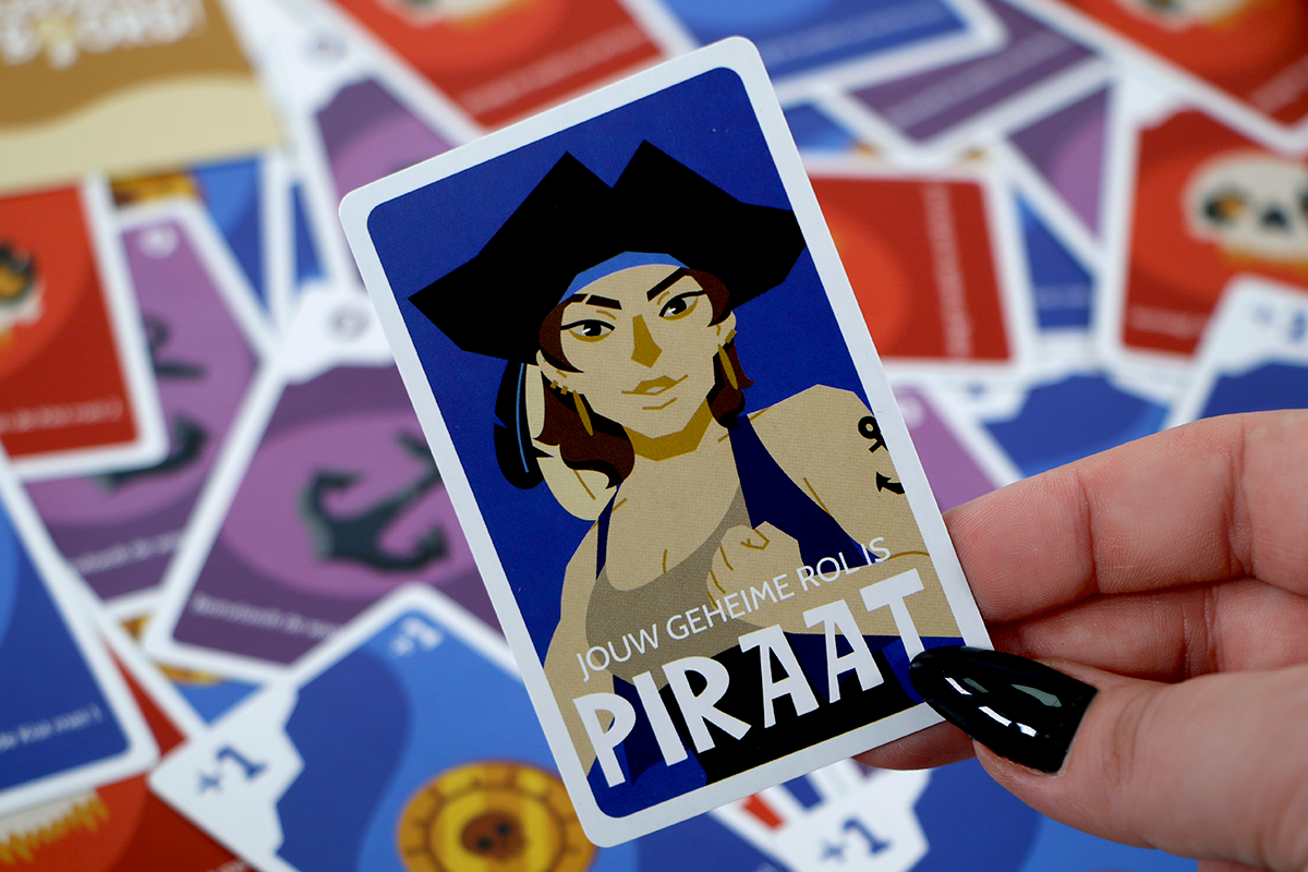 Hand die een kaart vasthoudt met een piratenillustratie en de tekst 'Jouw geheime rol is Piraat', omringd door andere kleurrijke kaarten.