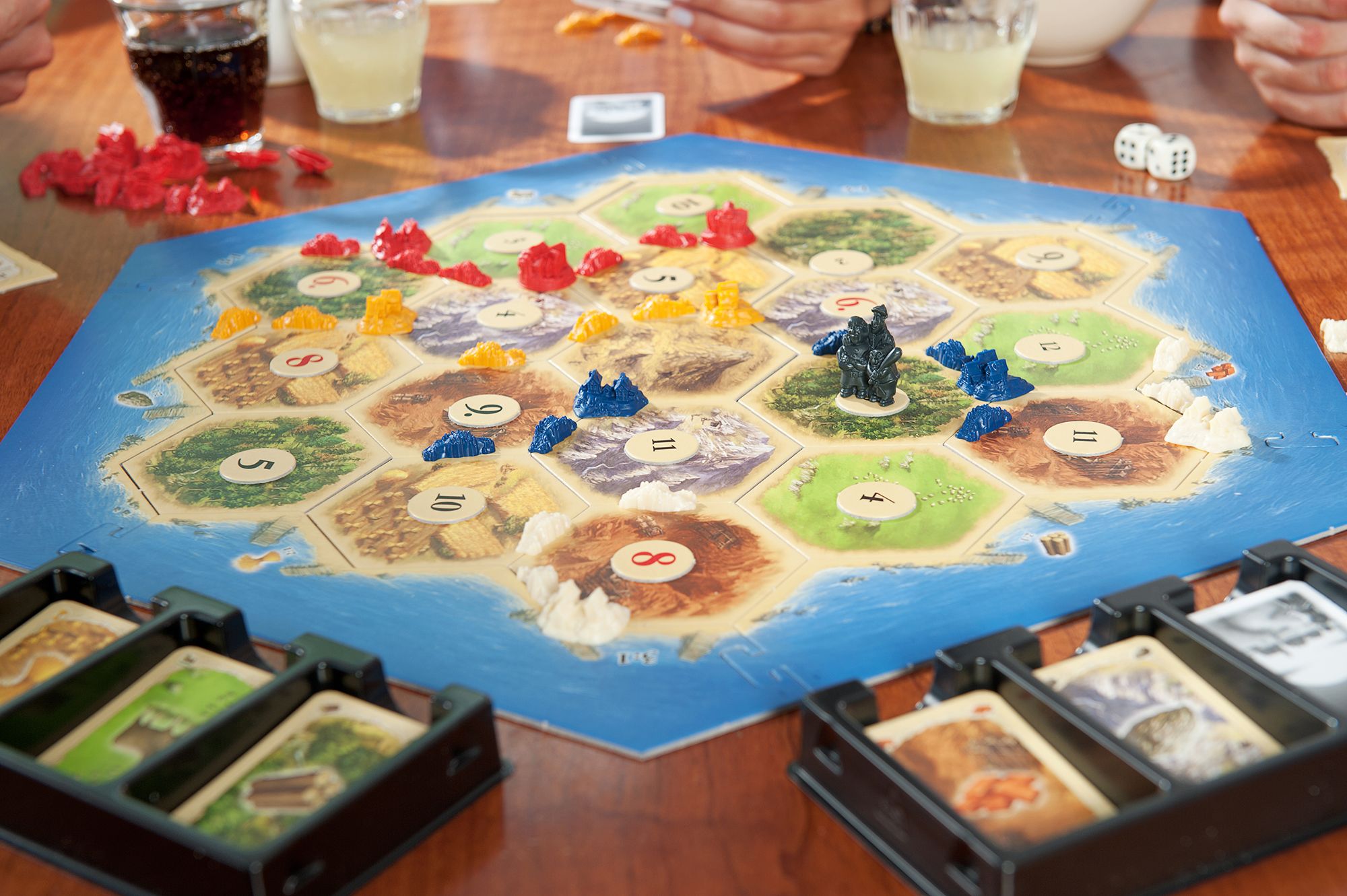 Bordspel CATAN met een opstelling van zeshoekige tegels, kleurrijke speelstukken, kaarten en dobbelstenen op een houten tafel. Handen van spelers zijn in de buurt zichtbaar.