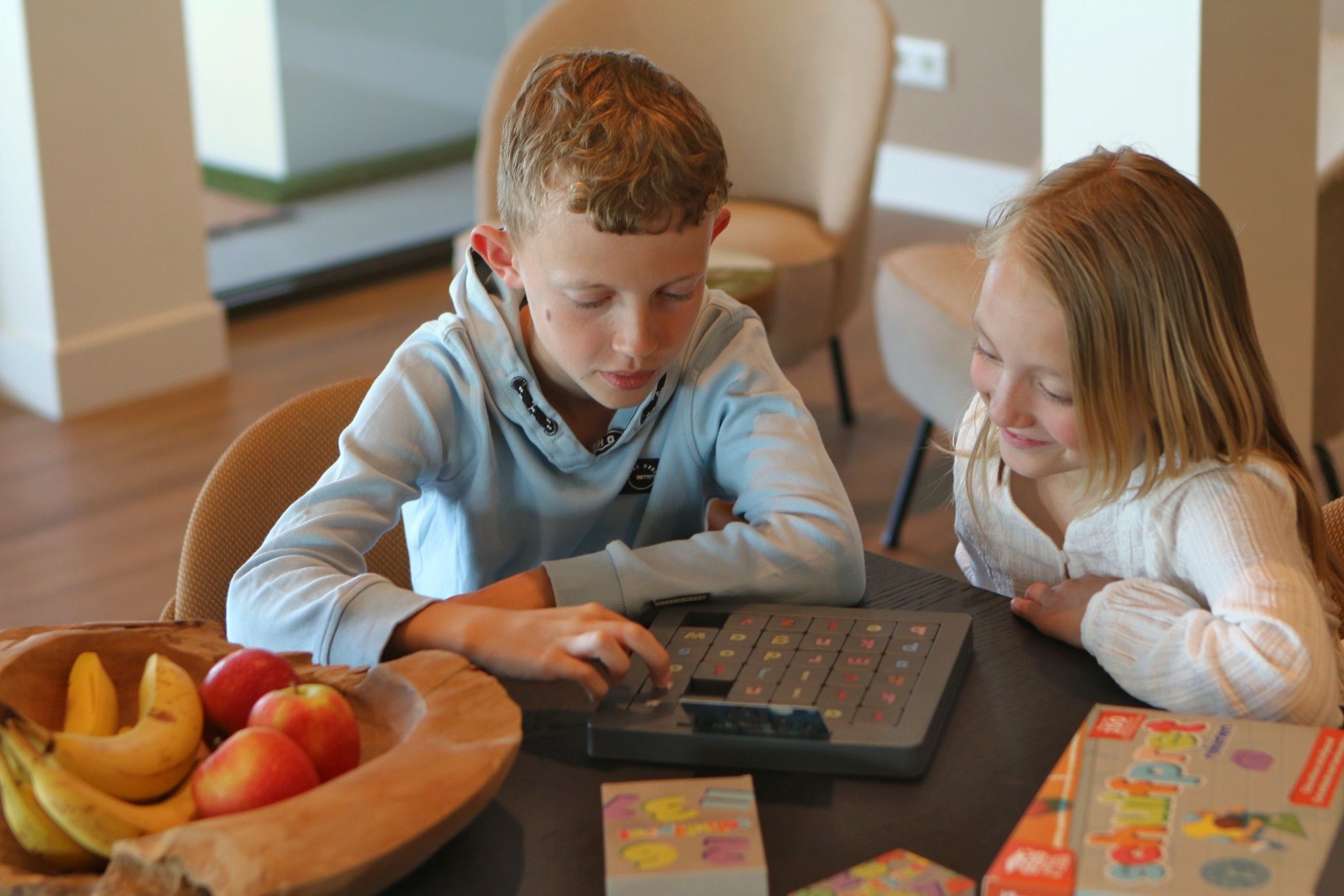 Twee kinderen zitten aan een tafel en spelen Schuifpret. Een schaal met fruit, met bananen en appels, staat ernaast.