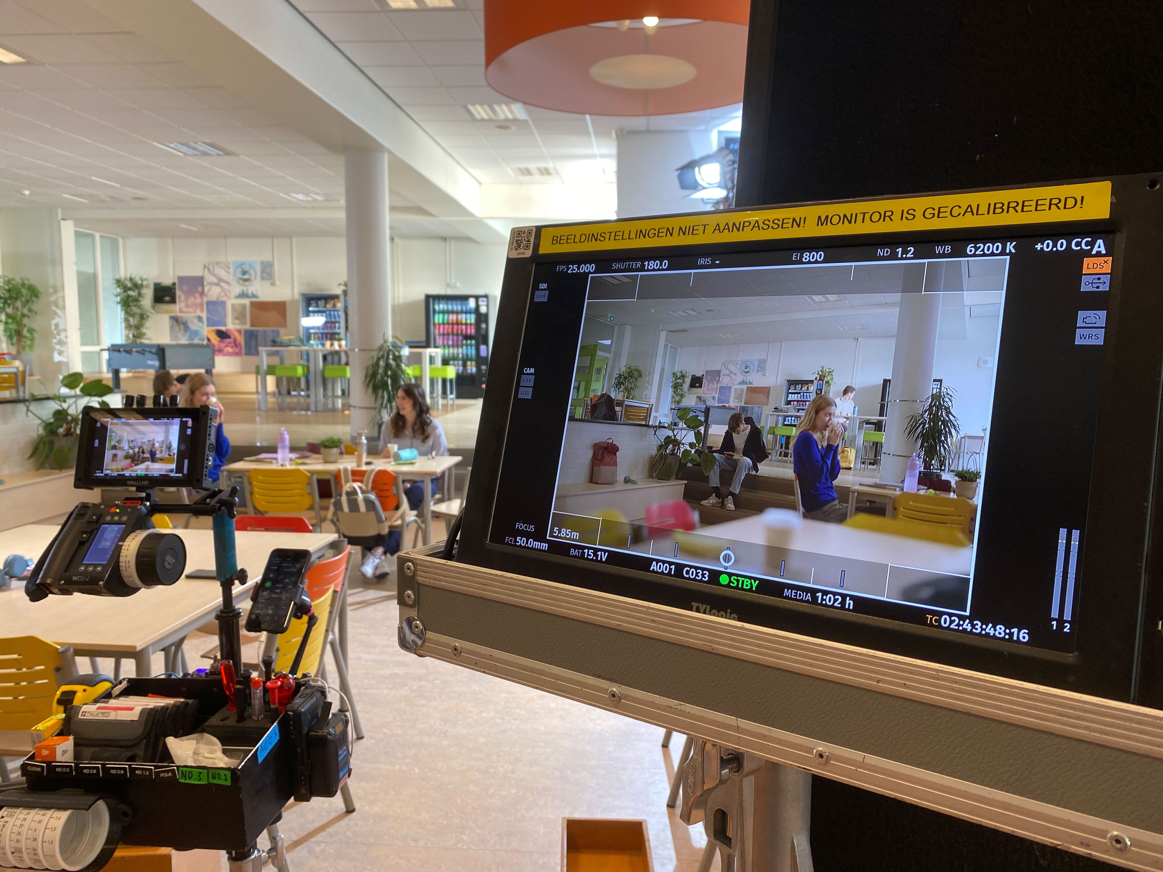 Filmset 999 Games commercial met een camera en een monitor die een scène toont in een lichte ruimte met mensen aan tafels, omringd door apparatuur en belichting.