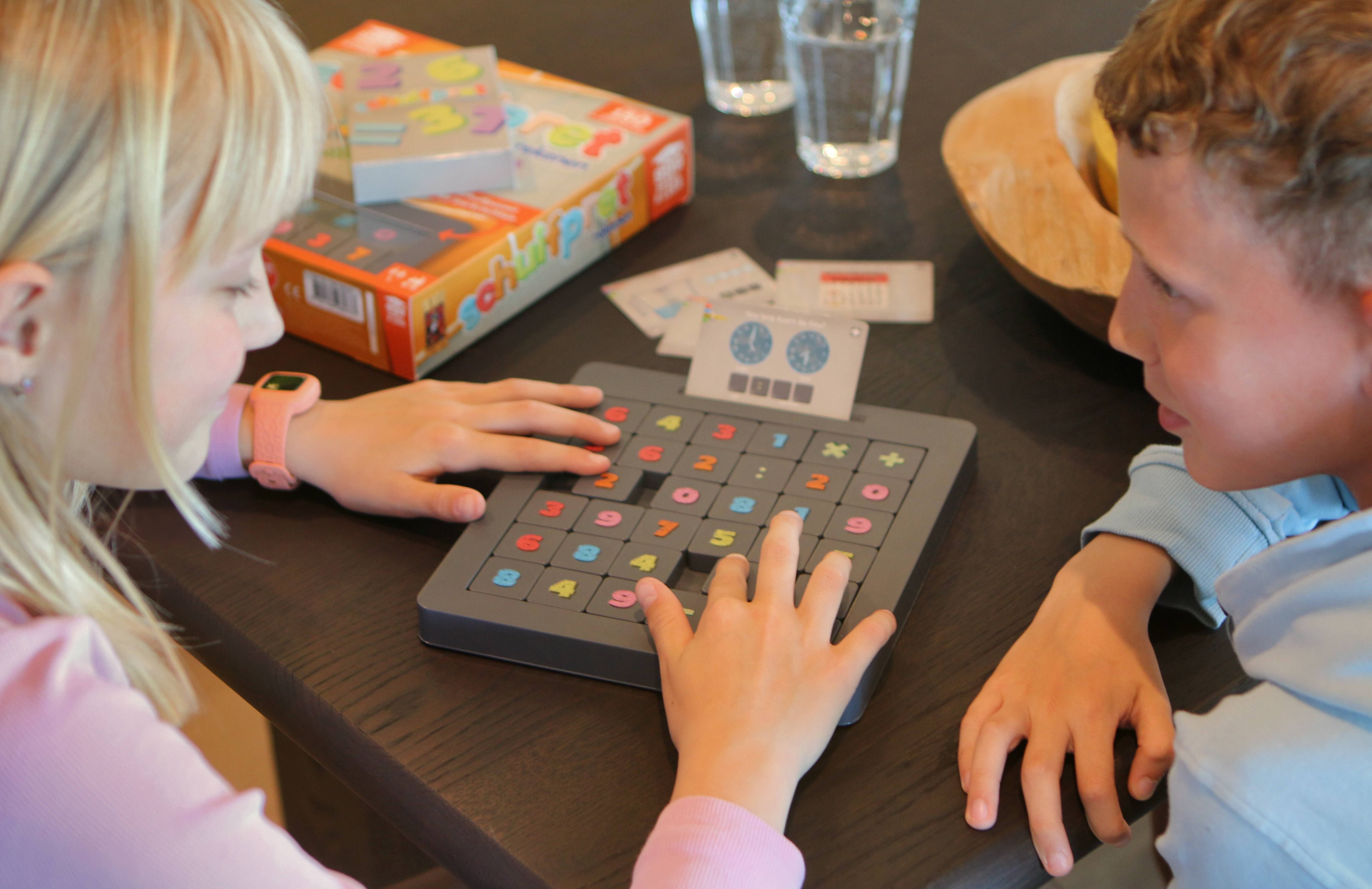 Twee kinderen spelen een bordspel met kleurrijke tegels op een tafel. Een speldoos en kaarten staan op de achtergrond.