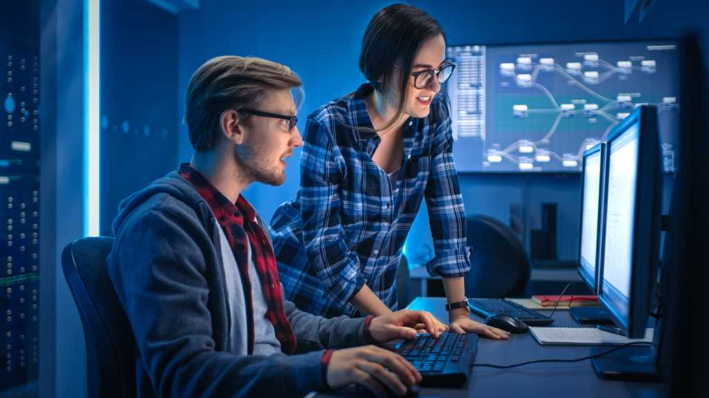 Two people in a blue-lit room working at computers, with data flow charts displayed on monitors in the background.