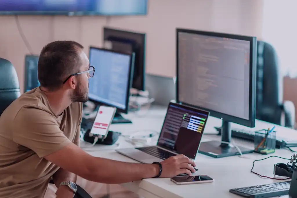 A person wearing glasses is working at a desk with multiple computer screens displaying code, using a laptop and a smartphone is nearby.