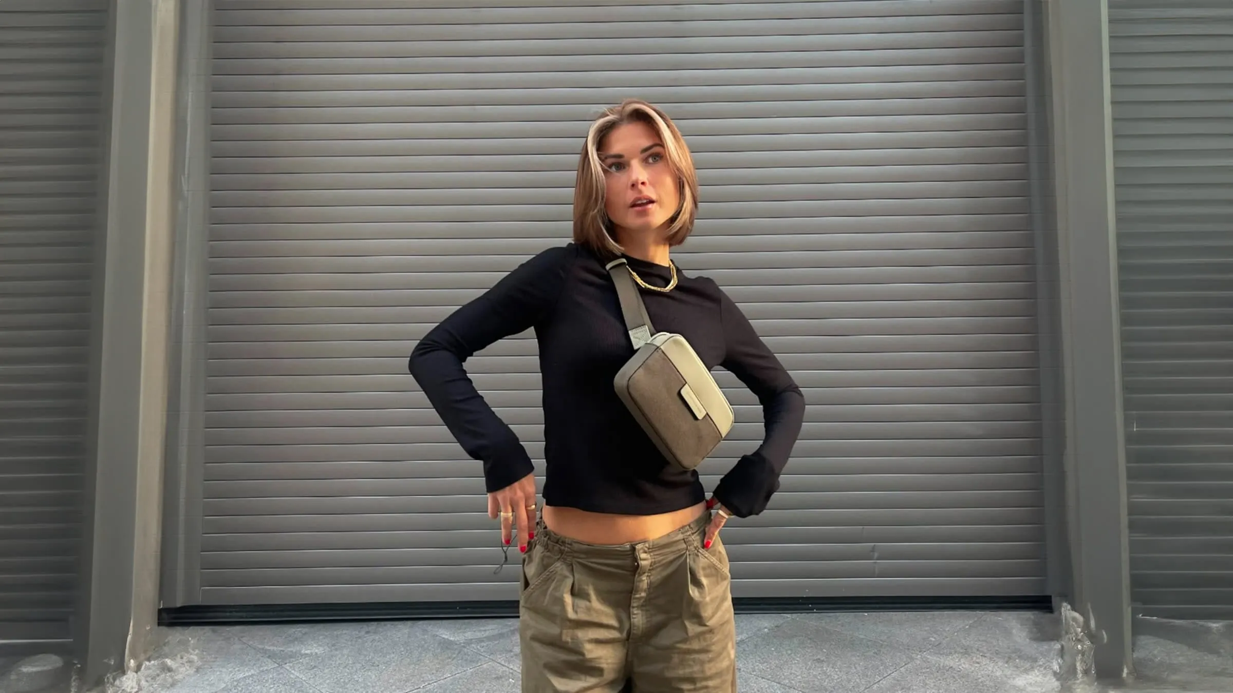 Frau steht vor grauem Rolltor mit schwarzem Oberteil und Umhängetasche.
