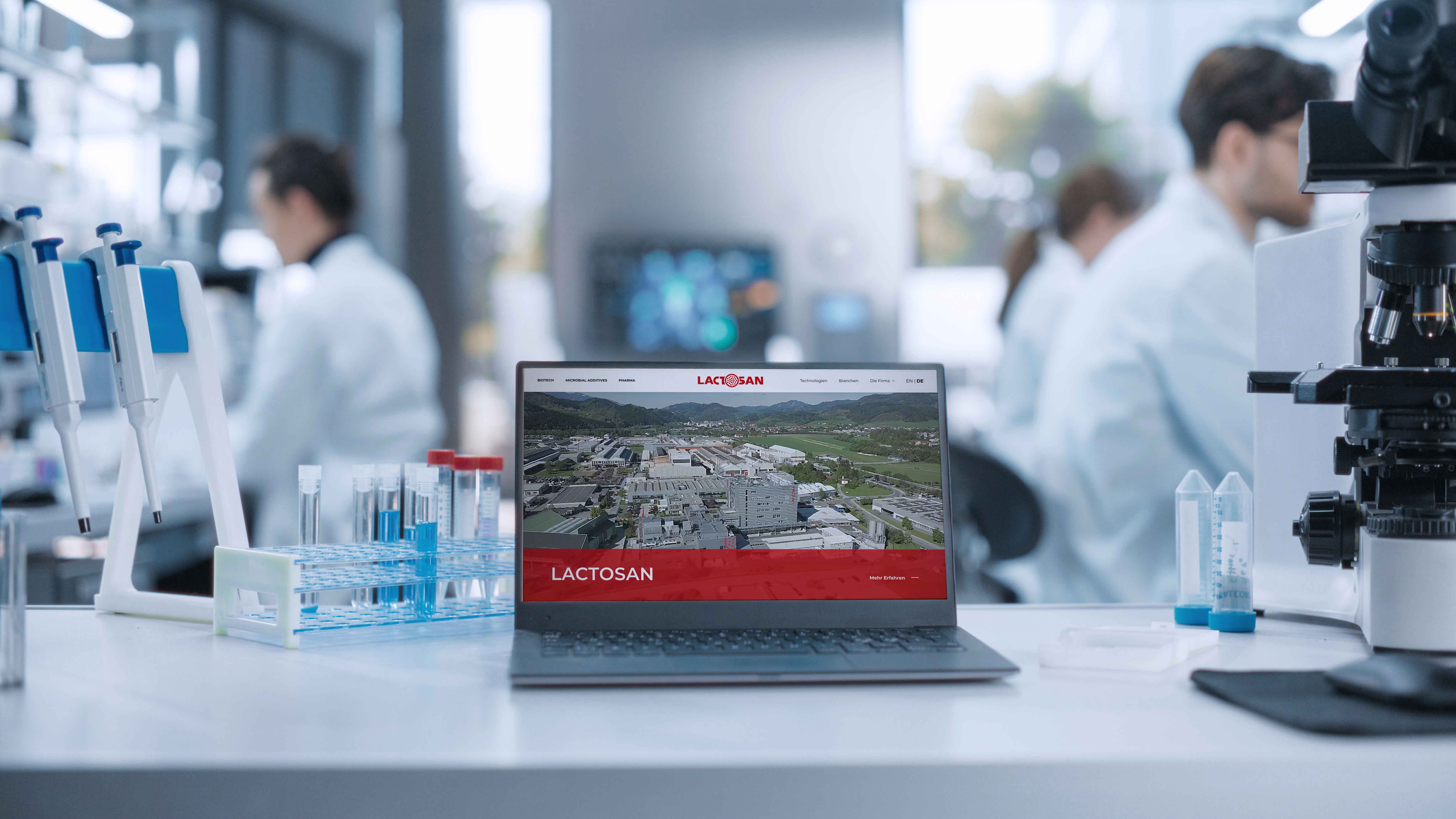Laptop displaying a website on a lab table with test tubes and a microscope; scientists work in the blurred background.