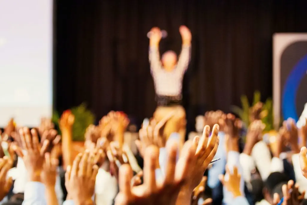 a mass of raised hands, in the background a person demonstrating the movement
