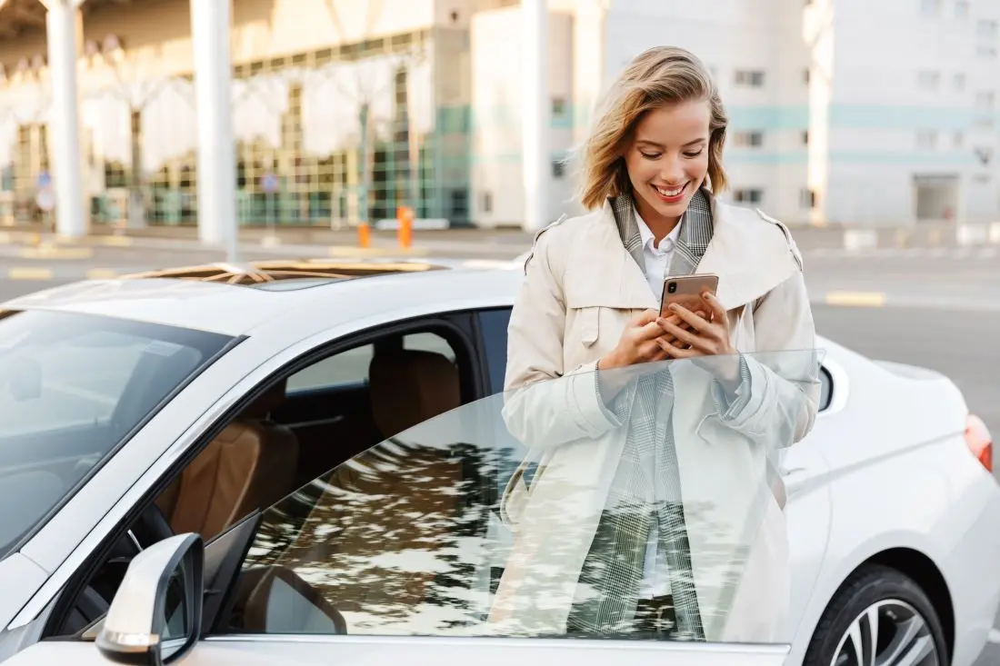 Frau steht in der offenen Autotür und blickt lächelnd auf ihr Smartphone.