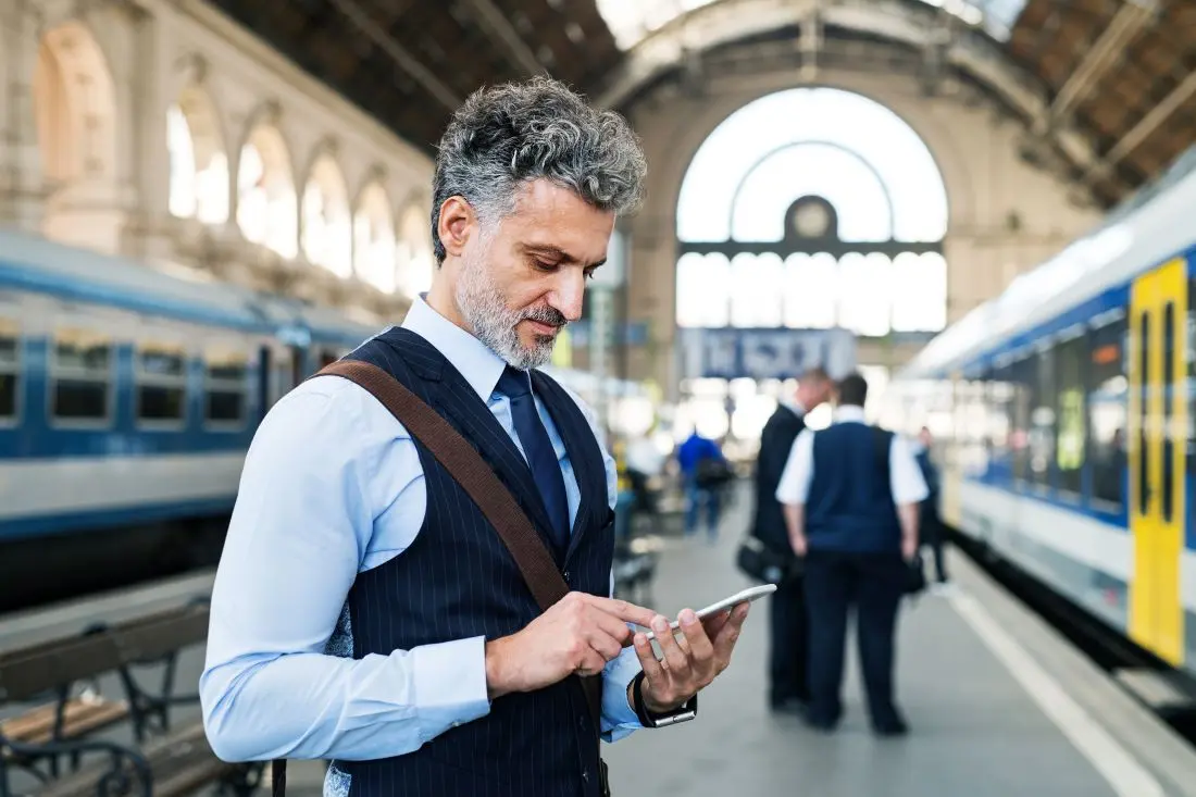 Mann in Businesskleidung steht am Bahnsteig und schaut auf sein Handy.