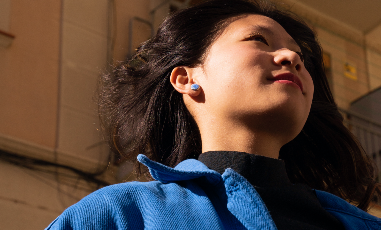 Woman in a blue jacket, looking upwards, with wind-blown hair, stands in sunlight against an urban background.