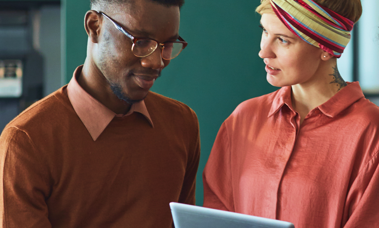 Two people in casual attire discussing something while looking at a laptop screen. One is a man wearing glasses, and the other is a woman with a headband.