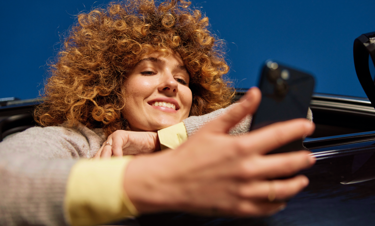 A person with curly hair smiles while looking at a smartphone, leaning out of a car window against a clear blue sky.