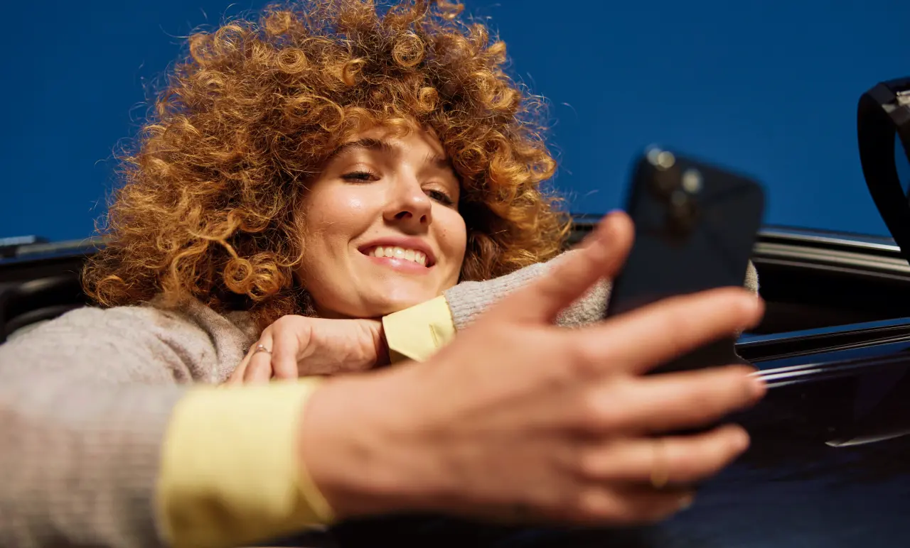 A person with curly hair smiles while looking at a smartphone, leaning out of a car window against a clear blue sky.