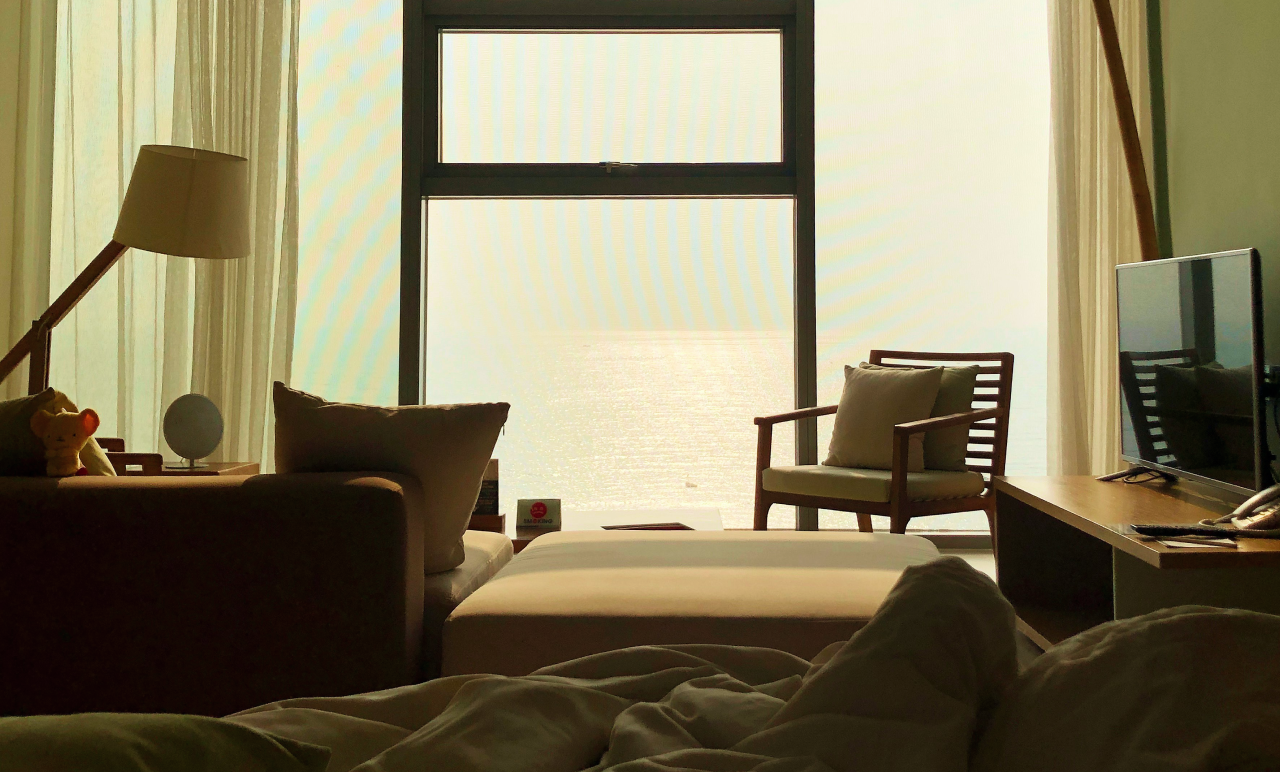 Cozy room with a bed, sofa, and chair by a large window overlooking a bright, hazy view. A TV and lamp are on the sides.