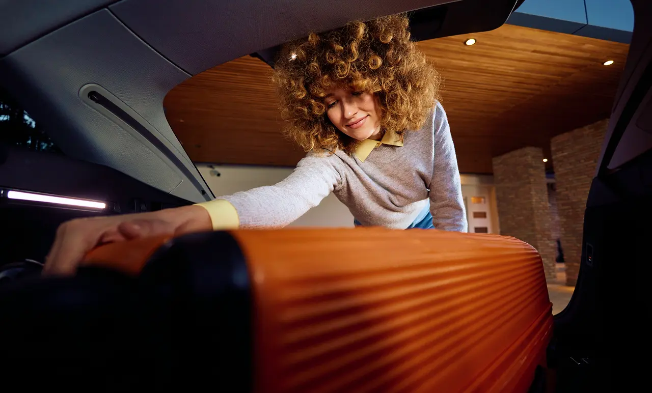 Woman with curly hair placing an orange suitcase into a car trunk, smiling. Indoor setting with wooden ceiling visible.