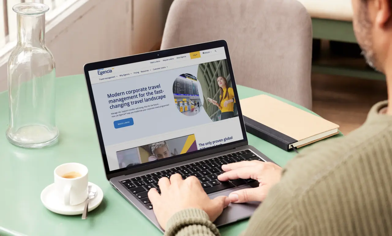 Egencia homepage shown on a laptop screen. A man is typing on the laptop keyboard and has a cup of coffee next to him