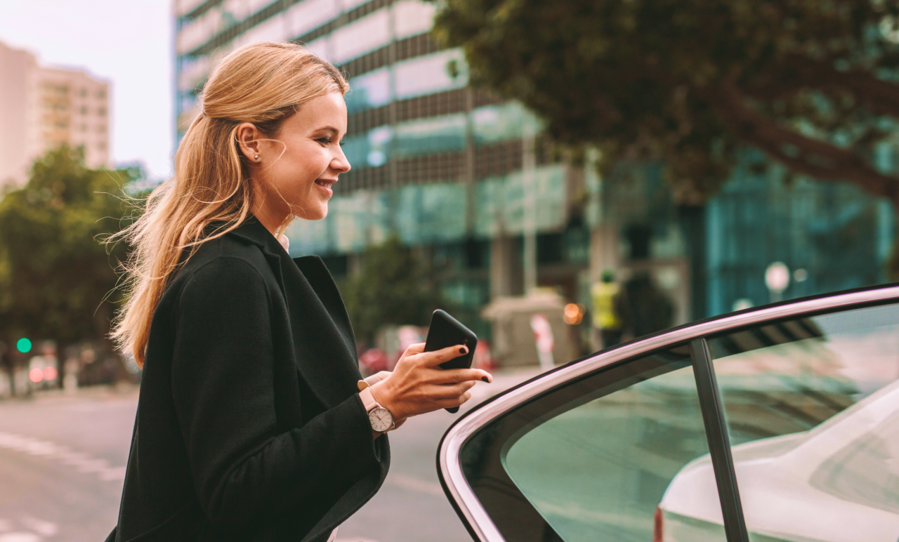 Smiling woman with long blonde hair enters a car on a city street, holding a smartphone, wearing a black coat. Urban buildings in the background.