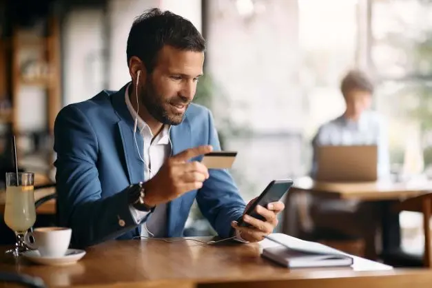 Mann im Anzug sitzt im Café und hält eine Kreditkarte und sein Smartphone in der Hand.