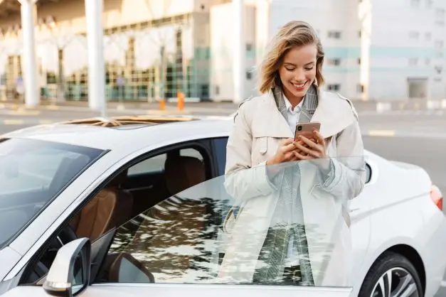 Frau steht in der offenen Autotür und blickt lächelnd auf ihr Smartphone.