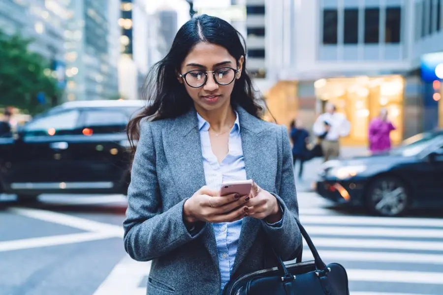 Frau in grauem Blazer und mit Brille steht auf einem Gehweg in der Stadt und schaut aufs Handy.