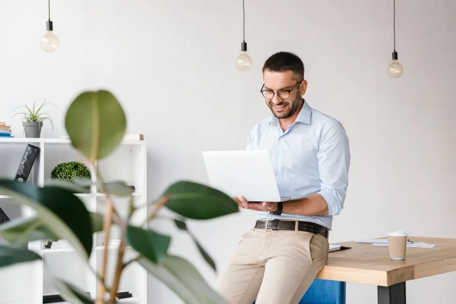 Mann in Businesskleidung lehnt sich mit einem Laptop in der Hand an einen Tisch.