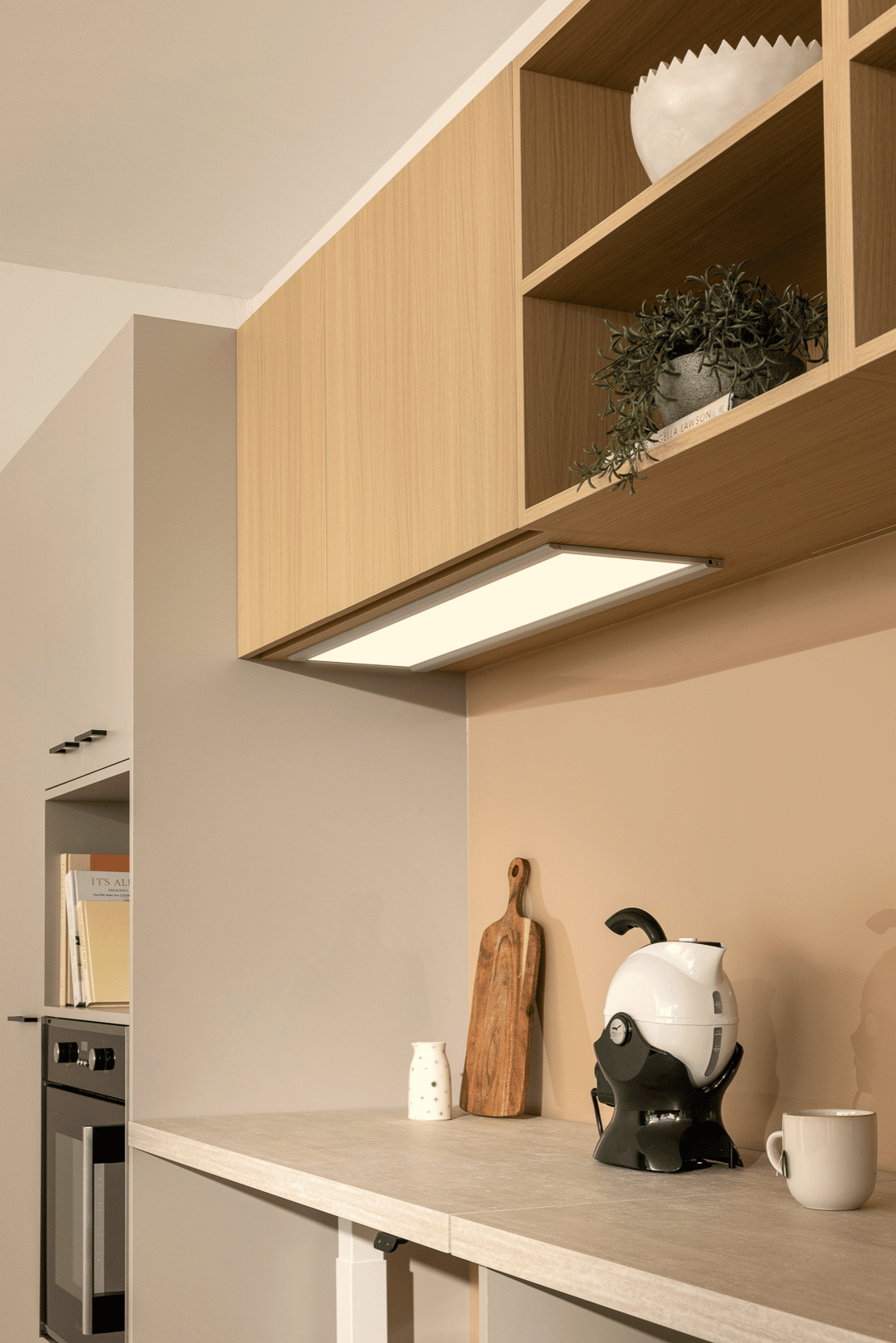 Modern kitchen with light wooden cabinets, oven, white kettle, mug, and rustic cutting board on a light countertop. Decorative plants above.