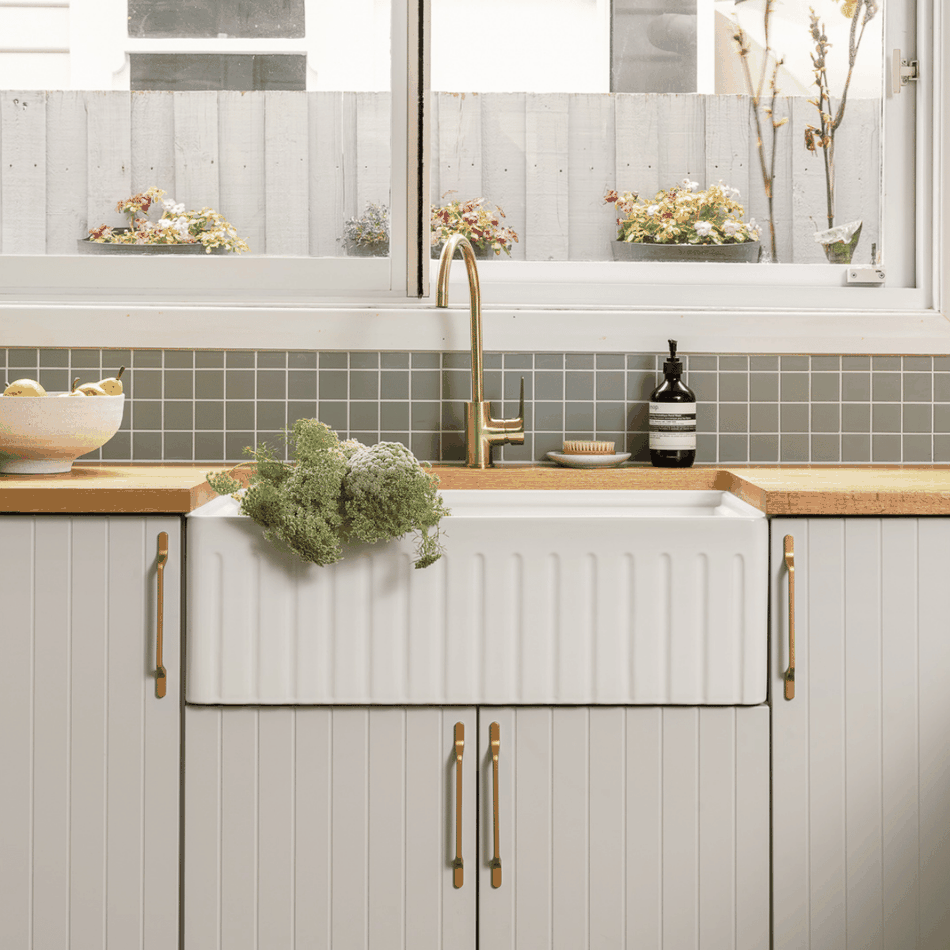 Farmhouse kitchen sink with gold faucet, wooden counter, and vase of flowers next to window with flower pots outside.