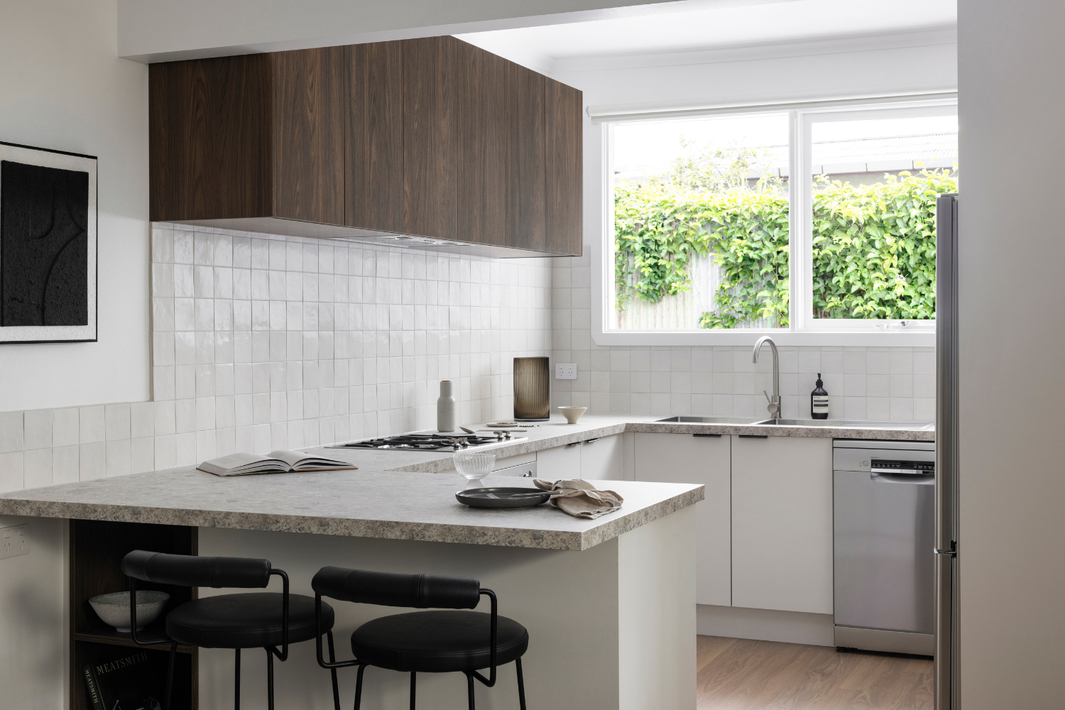 Modern kitchen with white cabinets, light benchtop, dark wood accents, breakfast bar with stools, and a window overlooking greenery.