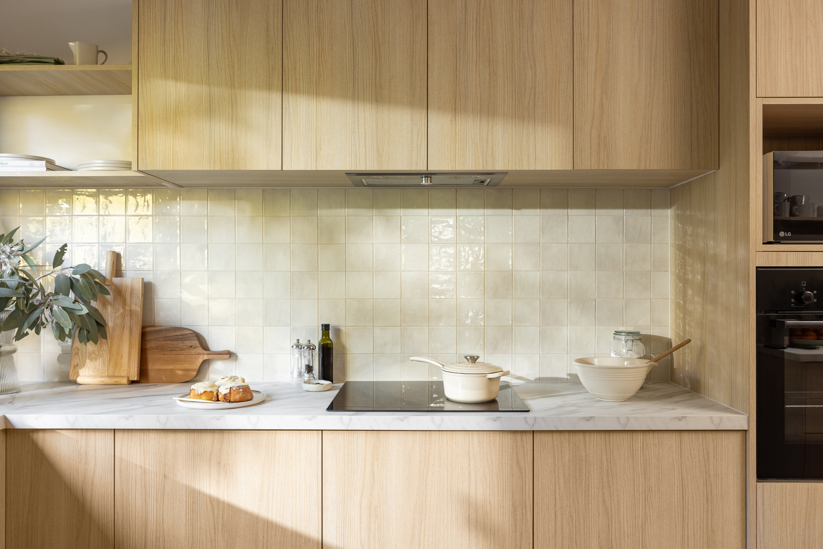 Modern kitchen with light wood cabinets, stone-look benchtops, cutting boards, olive oil bottle, and cooking utensils. Sunlight casts soft shadows.