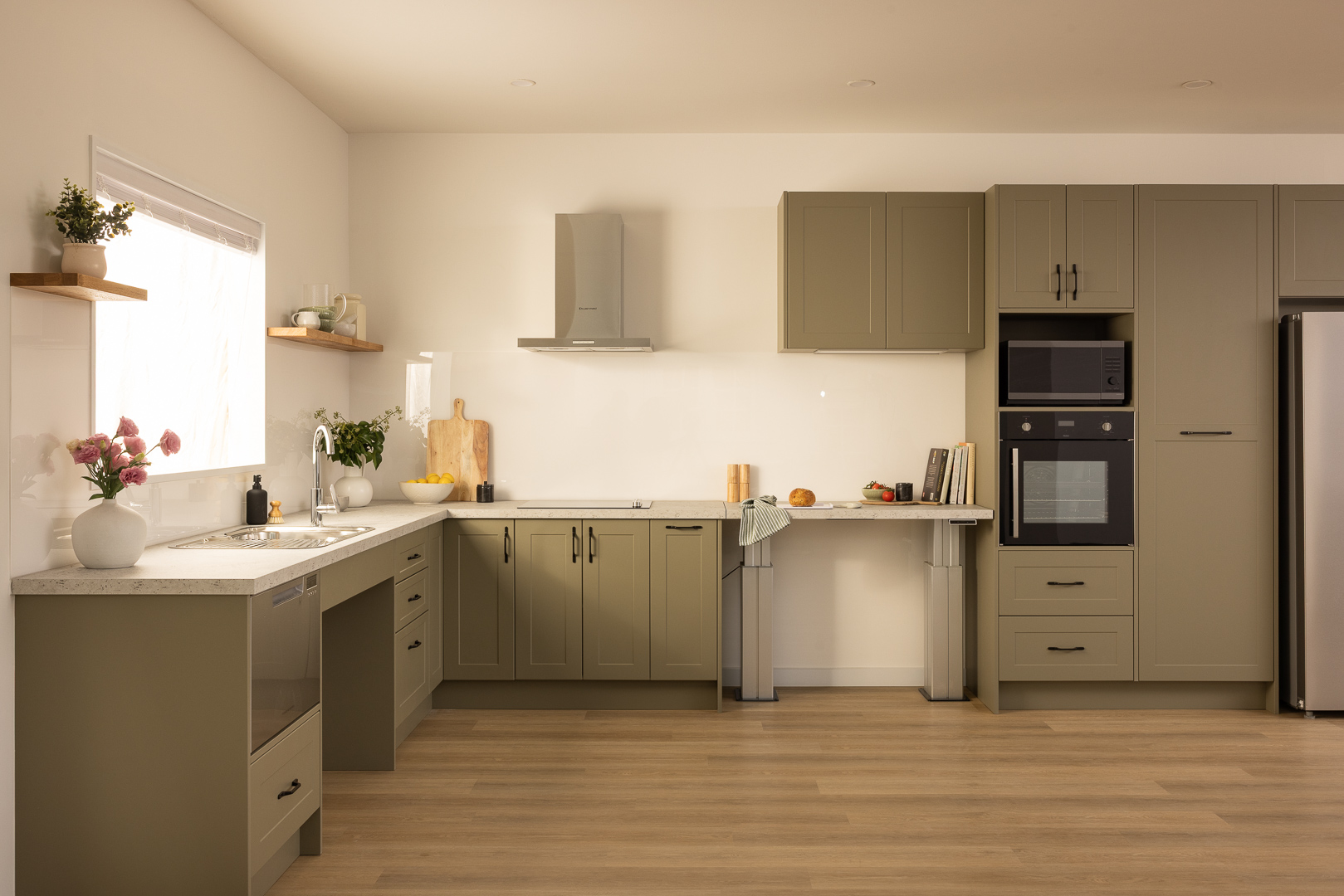 Modern kitchen with olive green cabinets, stainless steel appliances, and wooden shelves on a light wood floor. Decor includes plants and kitchenware.