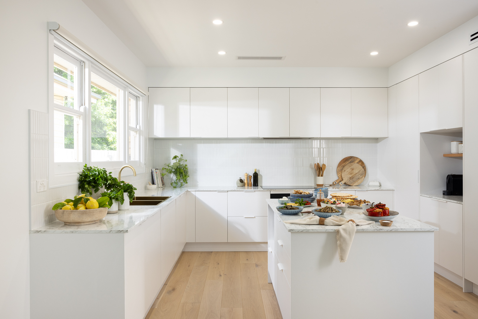Bright modern kitchen with white cabinets, light wood flooring, and a central island. Fresh produce and dishes are arranged on the countertops.