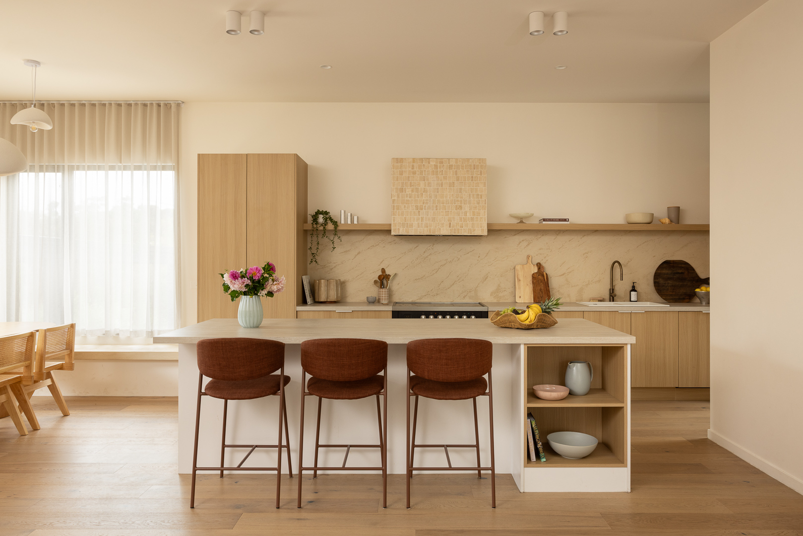 Coastak kitchen with wooden cabinets, marble backsplash, island with brown chairs, and a vase of flowers. Light wood flooring and dining area visible.
