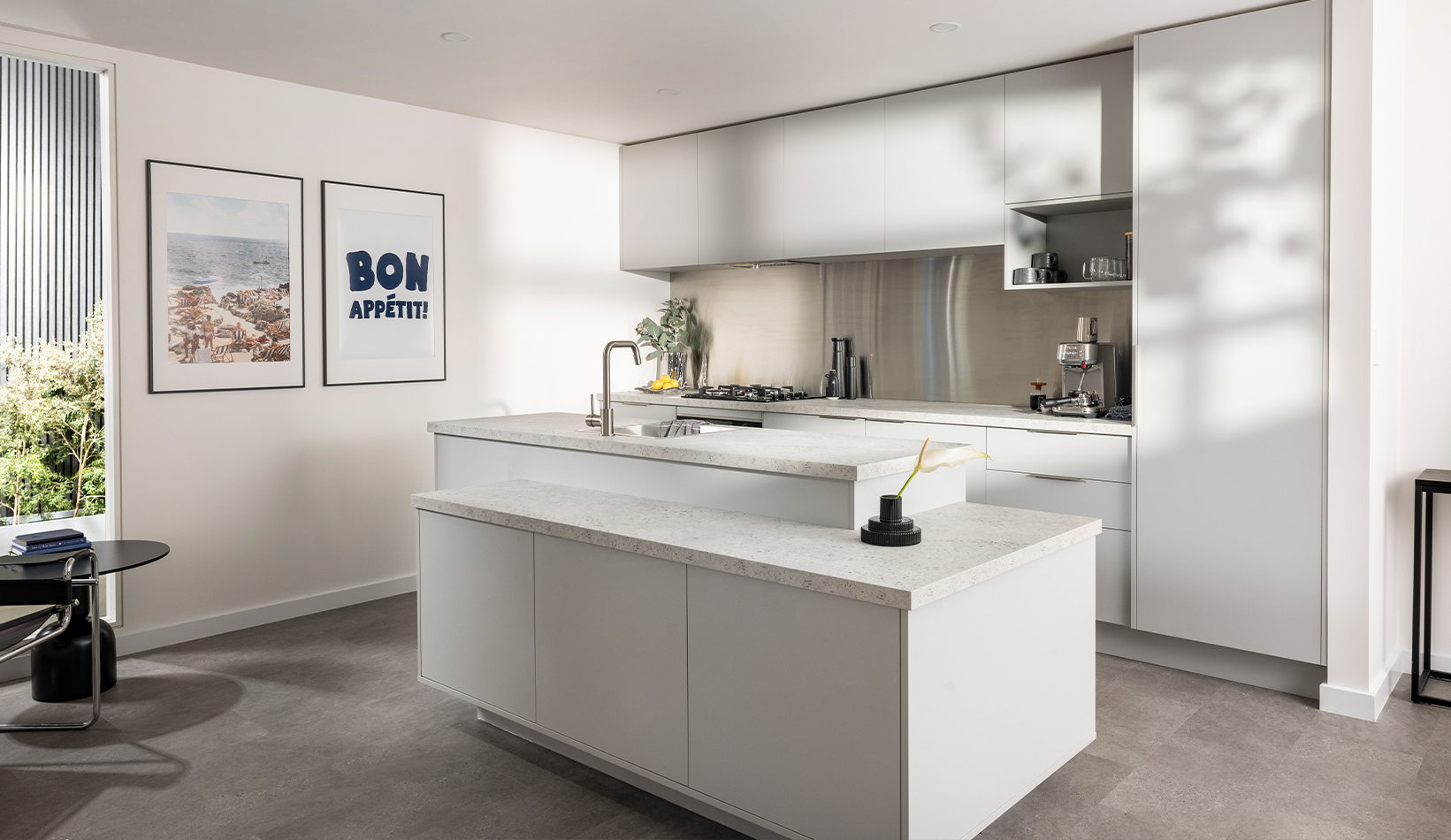 An industrial kitchen with grey cabinets, a central island benchtop, and minimalist decor. Sunlight streams through a window, illuminating the space.