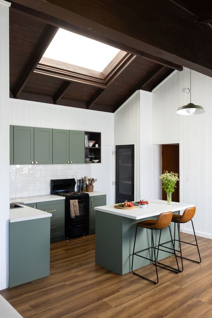 Modern kitchen with green cabinets, skylight, and wooden elements. Features an island with stools, flowers, and fruit, under pendant lighting.