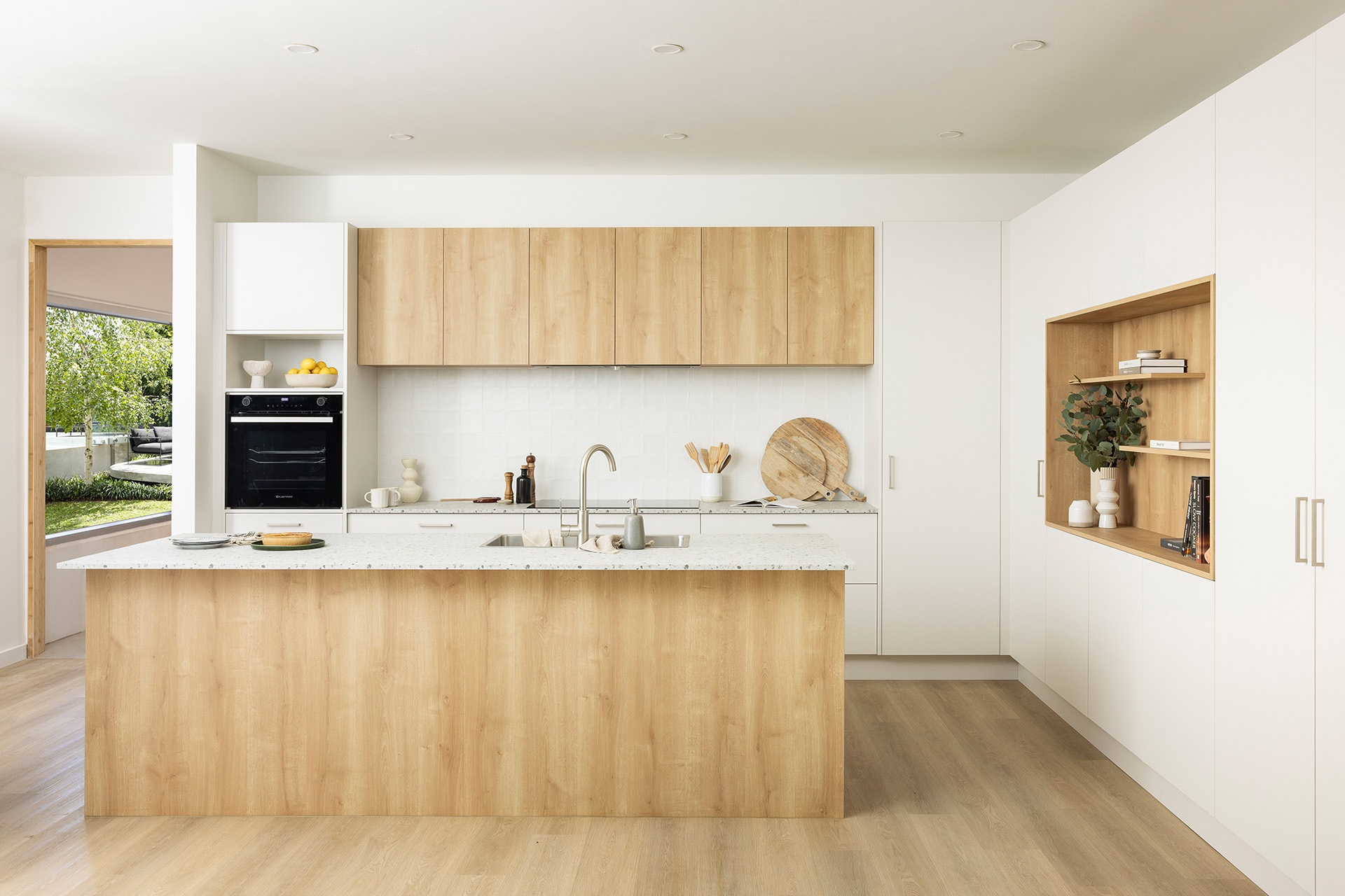 Modern kitchen with wooden cabinets, white countertops, island with sink, built-in oven, and open shelving, overlooking a garden through a sliding door.