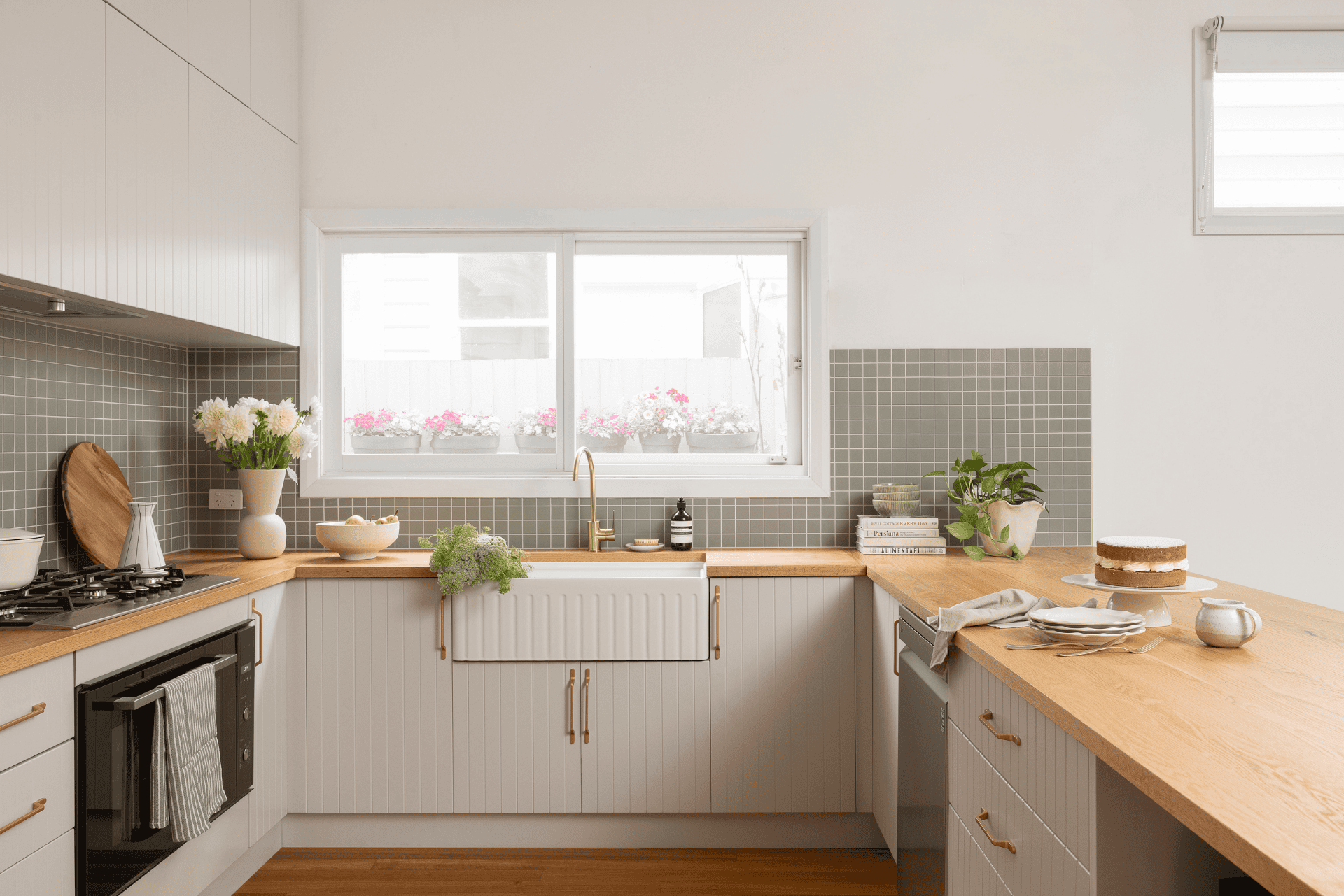 Bright kitchen with a farmhouse sink, timber countertops, white cabinets, and a large window. Accents include plants, dishes, and a cake.