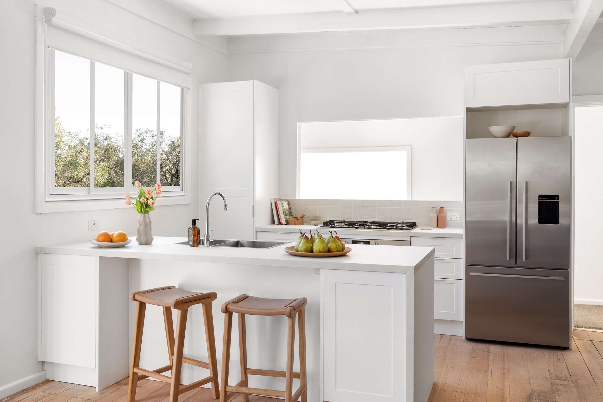 Modern white kitchen with a large island bench, stainless steel fridge, and a vase of flowers beside a bowl of pears.
