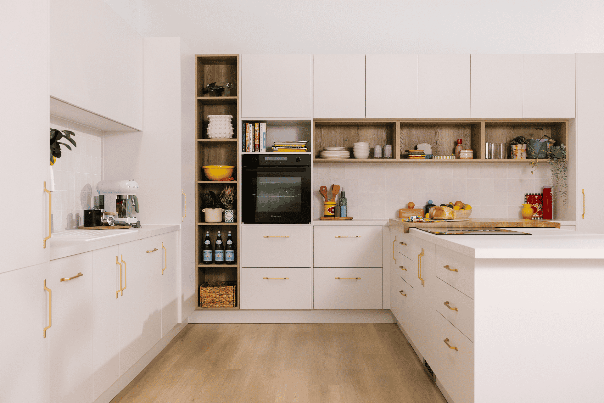 Modern kitchen with white cabinets, gold handles, and an island. Open shelves display kitchenware. Light wood floor and integrated oven.