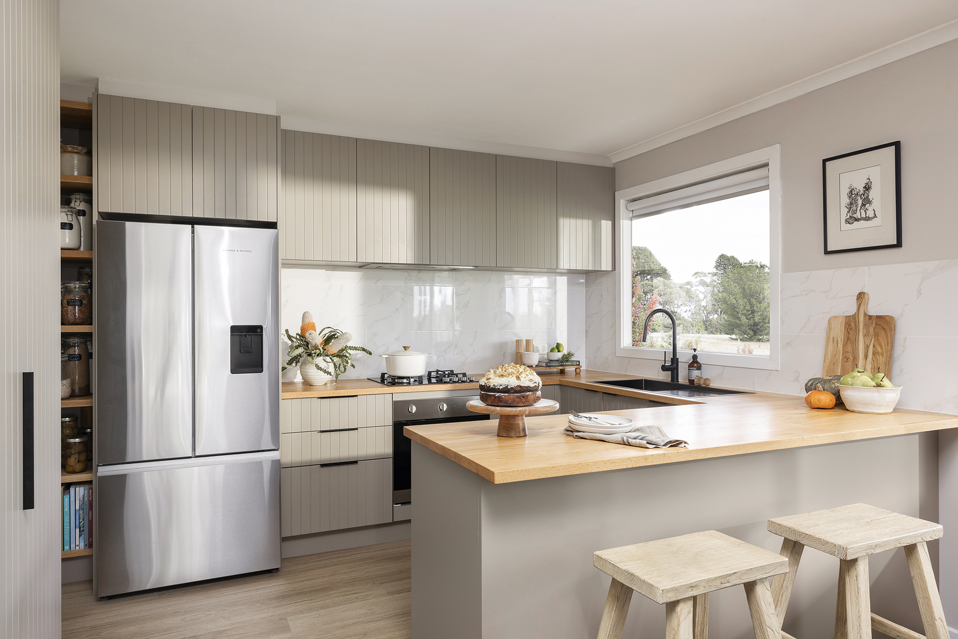 Modern kitchen with light wood cabinetry, stainless steel appliances, a wooden countertop, and bar stools. A large window brings in natural light.