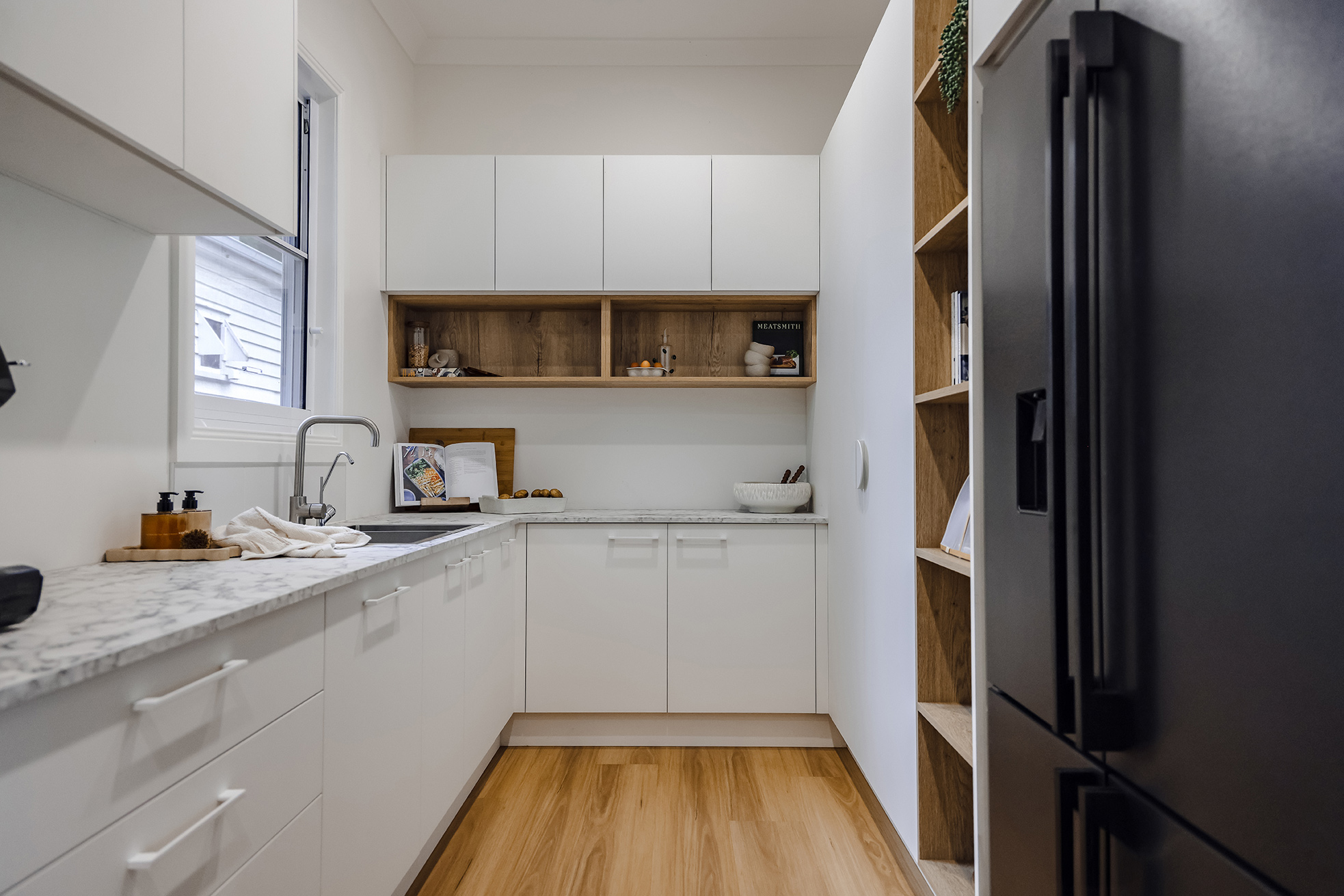 Modern white galley kitchen with timber flooring, white cabinetry, marble-look benchtops and open timber shelving, featuring a large dark fridge on the right.