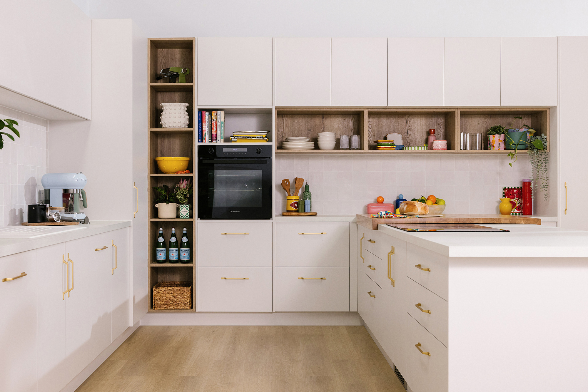 Modern kitchen with white cabinets, wooden shelves, under-counter oven, and an island. Decor includes books, plants, and colorful kitchenware.
