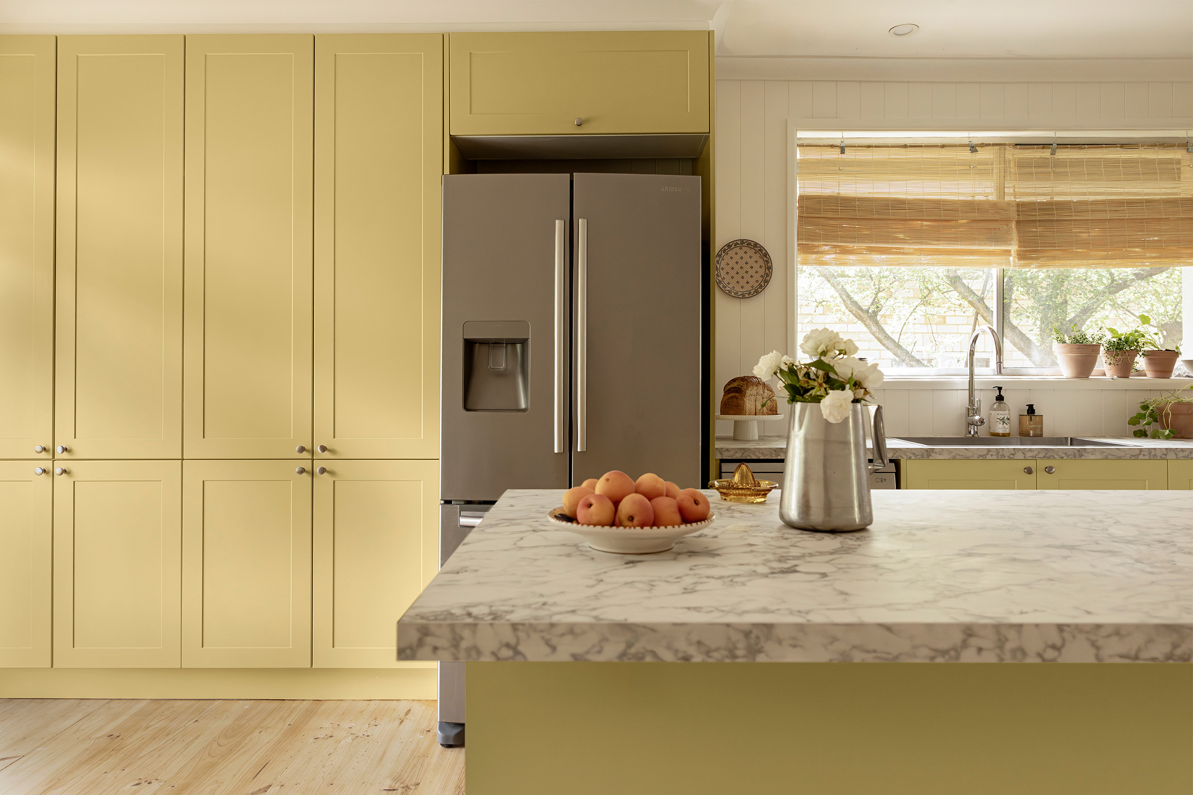 A country style kitchen with butter yellow cabinets and a stone-look countertop. A bowl of oranges and a vase of flowers on the island.