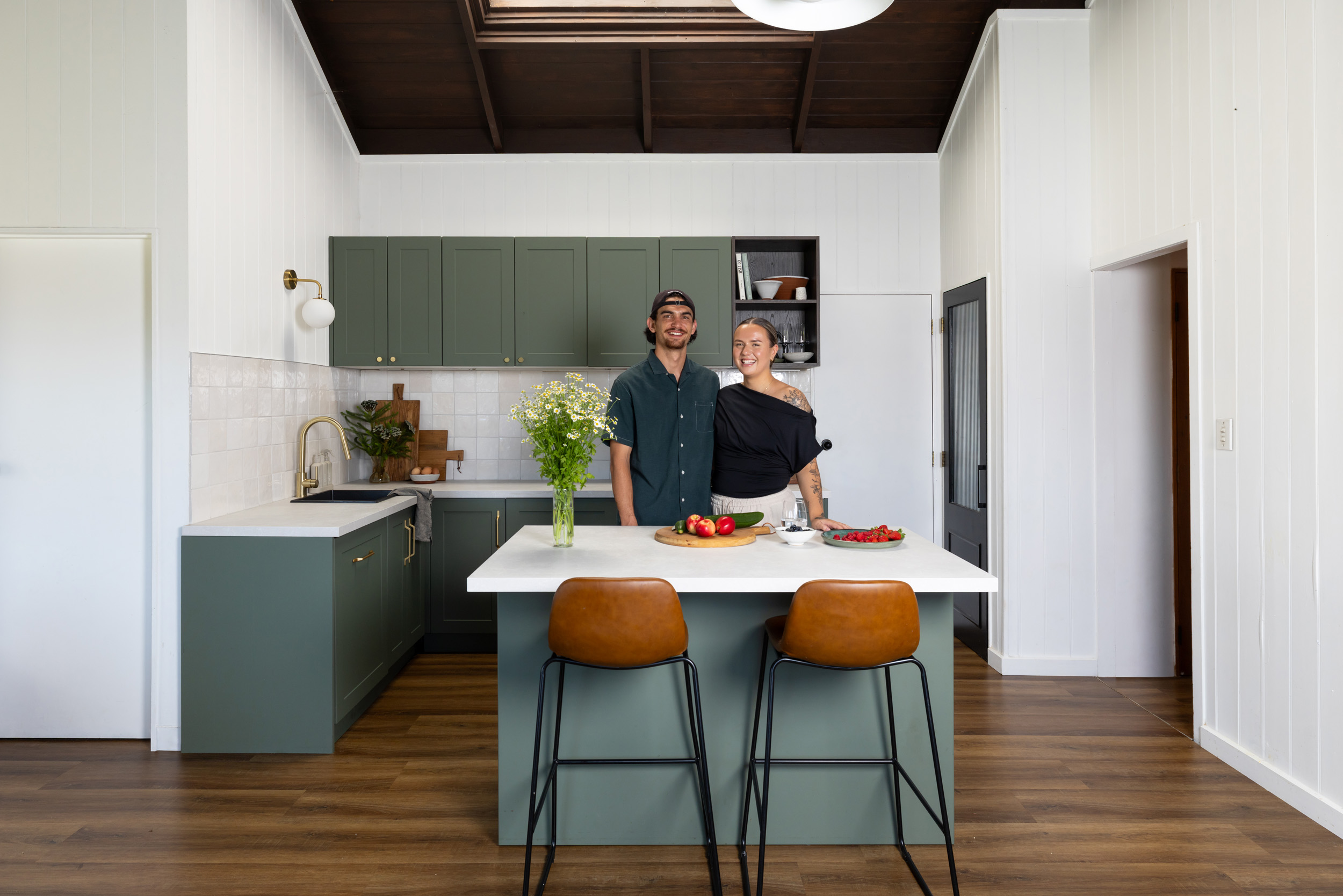 A smiling couple stands in a modern kitchen with green cabinets, a white island, flowers, and produce. Brown stools are placed at the island.