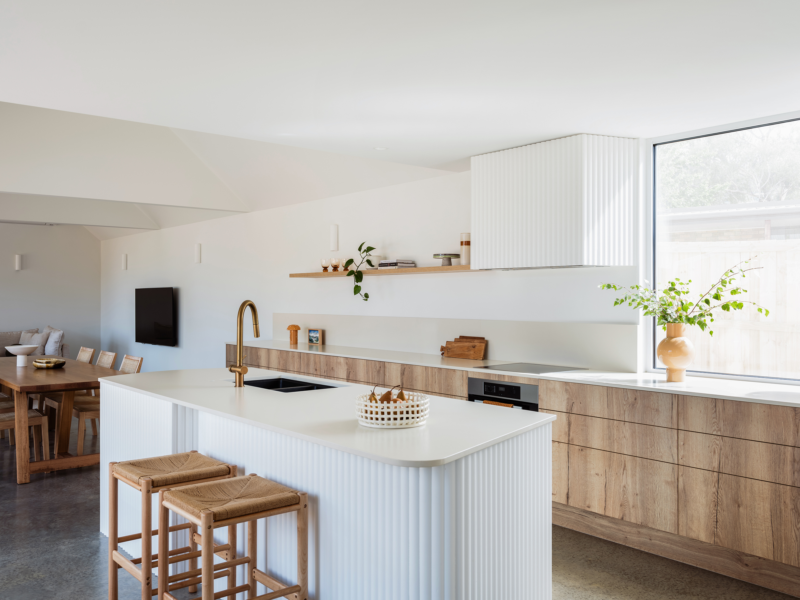 Scandi kitchen with a white island, wooden stools, open shelves, and a dining area. Large window allows natural light. Simple decor and plants.