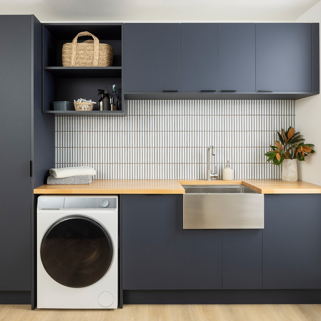 Modern laundry room with dark cabinets, wooden countertop, stainless steel farmhouse sink, washing machine, and decorative items on shelves.
