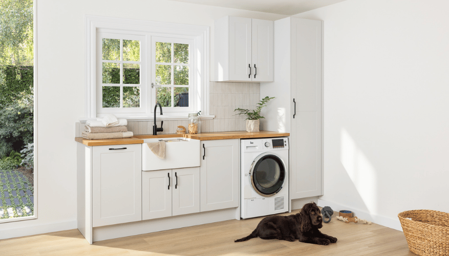 Bright laundry room with white cabinets, wood countertop, farmhouse sink, and a large window. A black dog rests on the wooden floor near a basket.