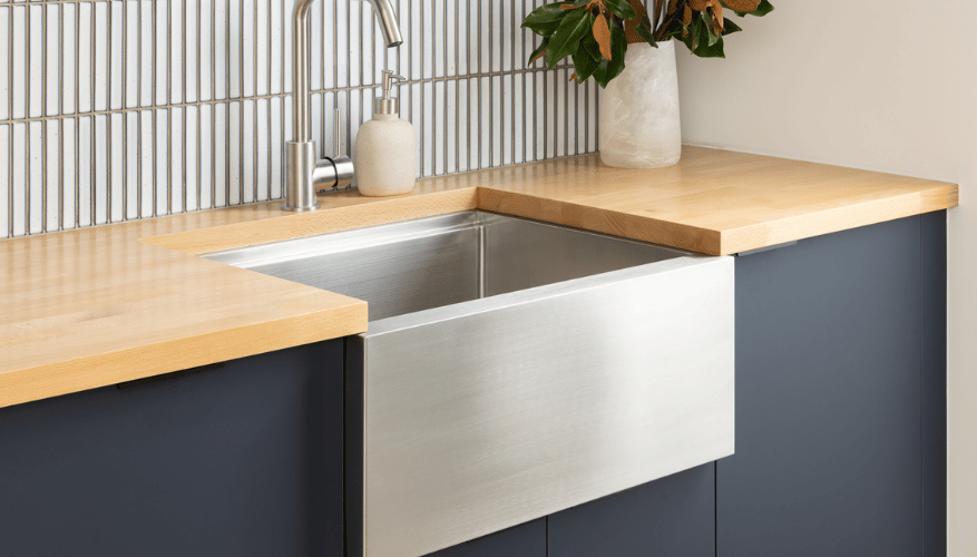 Modern kitchen corner with a stainless steel farmhouse sink, wooden countertops, gray cabinets, tiled backsplash, and a plant in a vase.