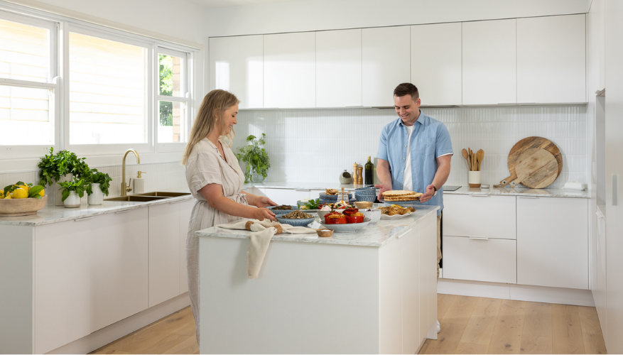 A couple preparing a meal in a bright, modern kitchen, with plants, fruits, and cutting boards on the counter, under natural light.