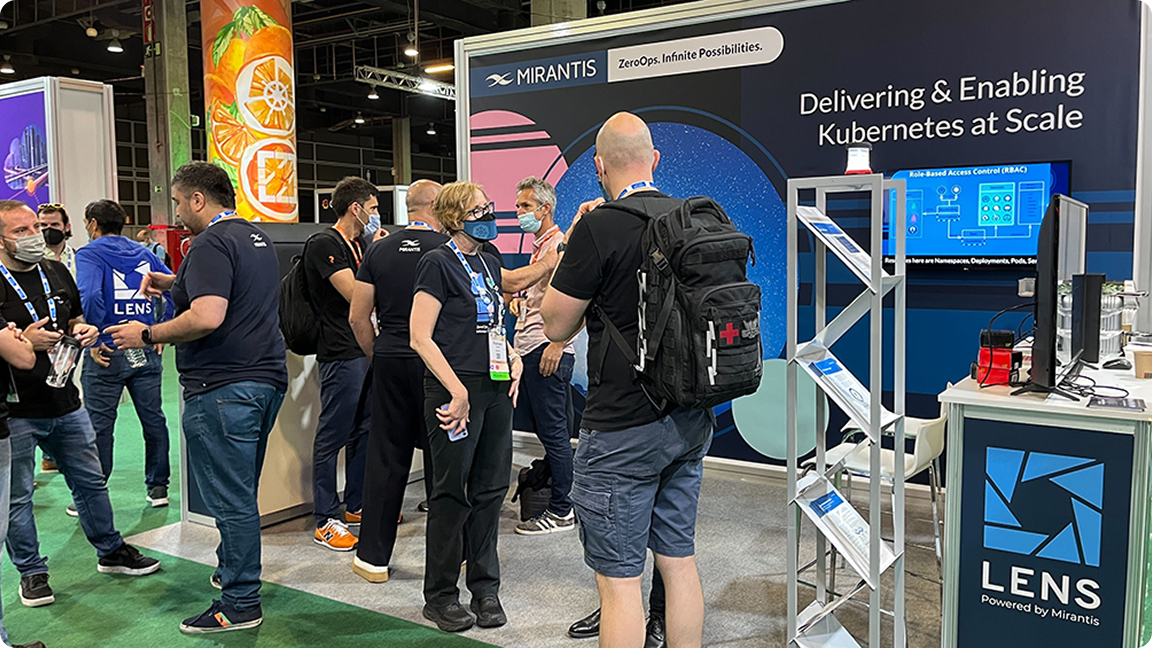 People gather at a Mirantis booth during a tech event, discussing Kubernetes with display screens and promotional materials around.
