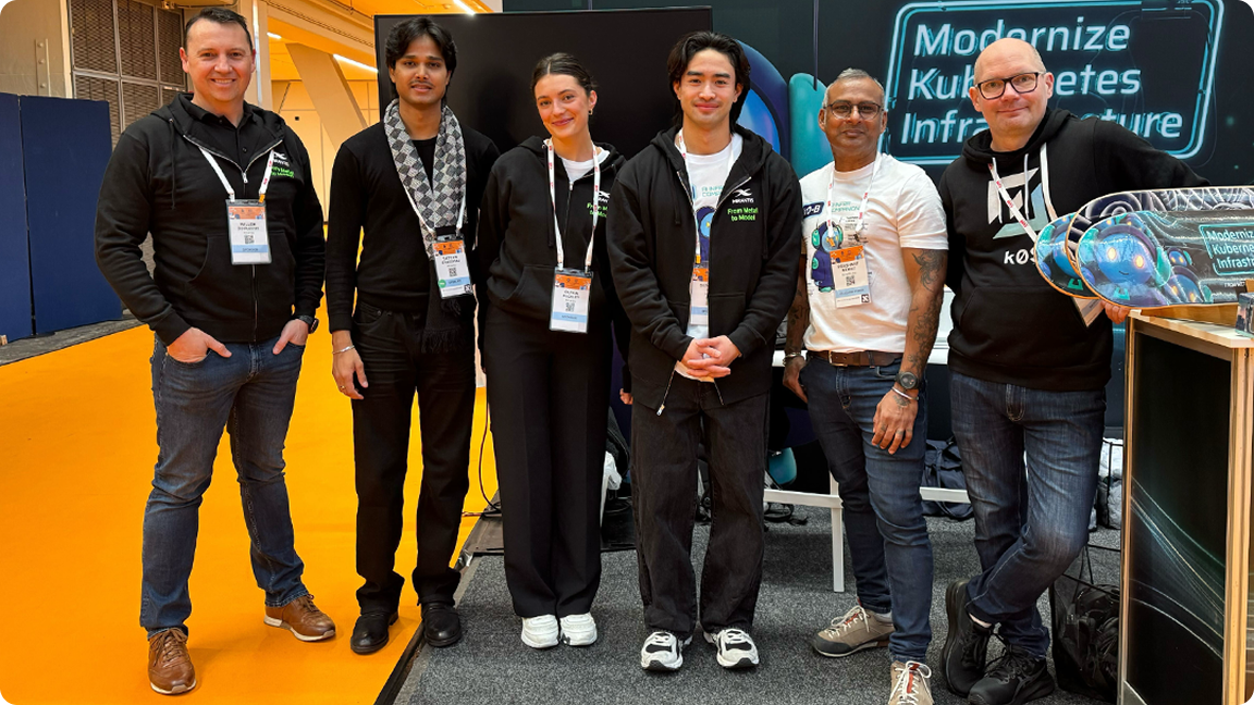 Six people standing together at a technology event, wearing lanyards and casual outfits, with a sign reading Modernize Kubernetes Infrastructure.
