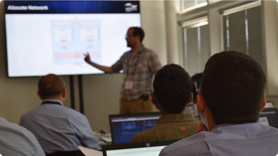A man presents a slide to an audience in a conference room. Attendees face the screen with laptops open, listening attentively.